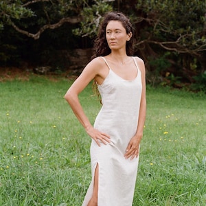 Puede incluir: Una mujer lleva un vestido blanco de lino con una abertura lateral. El vestido tiene un dise&ntilde;o sencillo, sin mangas y un ajuste holgado. Est&aacute; de pie en un campo de hierba con &aacute;rboles en el fondo.