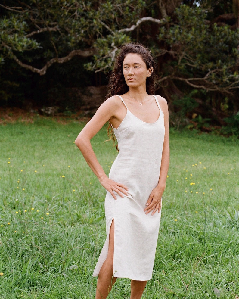 Puede incluir: Una mujer lleva un vestido blanco de lino con una abertura lateral. El vestido tiene un dise&ntilde;o sencillo, sin mangas y un ajuste holgado. Est&aacute; de pie en un campo de hierba con &aacute;rboles en el fondo.