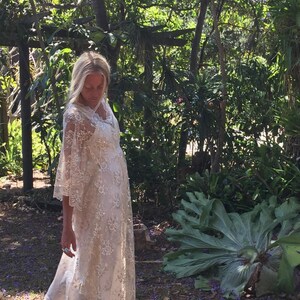K&ouml;nnte beinhalten: Eine Frau in einem wei&szlig;en Spitzenkleid mit Glocken&auml;rmeln steht in einer Waldlandschaft. Das Kleid hat ein zartes Blumenmuster und einen flie&szlig;enden Rock. Die Frau ist barfu&szlig; und hat langes blondes Haar.