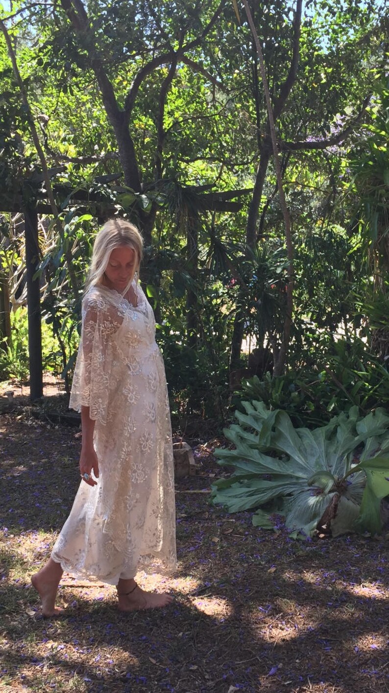 K&ouml;nnte beinhalten: Eine Frau in einem wei&szlig;en Spitzenkleid mit Glocken&auml;rmeln steht in einer Waldlandschaft. Das Kleid hat ein zartes Blumenmuster und einen flie&szlig;enden Rock. Die Frau ist barfu&szlig; und hat langes blondes Haar.