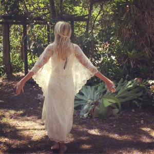 K&ouml;nnte beinhalten: Eine Frau in einem wei&szlig;en Spitzenkleid mit langen &Auml;rmeln und einem flie&szlig;enden Rock. Sie steht in einem Garten mit &uuml;ppigem Gr&uuml;n und Sonnenschein.