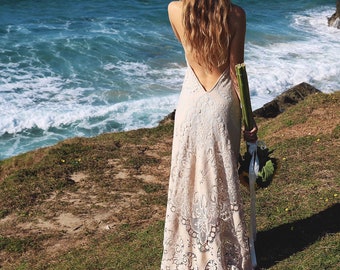May include: A woman wearing a white lace wedding dress with a low back and a floral crown stands on a grassy cliff overlooking the ocean.