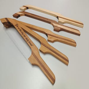 May include: Four wooden bread slicers with varying wood grain patterns. One slicer has the text "Give us this day our daily bread" engraved on the side. The slicers have a handle and a blade for cutting bread. The wood tones range from light to dark.