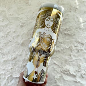 May include: White and gold insulated tumbler featuring a Wonder Woman design. The tumbler has a white base with a gold illustration of Wonder Woman and her emblem. The lid is light grey.