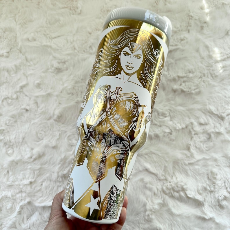 May include: White and gold insulated tumbler featuring a Wonder Woman design. The tumbler has a white base with a gold illustration of Wonder Woman and her emblem. The lid is light grey.