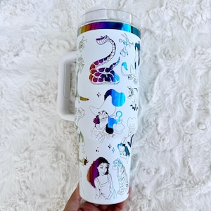 Puede incluir: Vaso blanco con asa y tapa transparente, adornado con ilustraciones de personajes de dibujos animados coloridos. El dise&ntilde;o incluye una banda iridiscente arco&iacute;ris en la parte superior. El vaso est&aacute; sostenido por una mano.