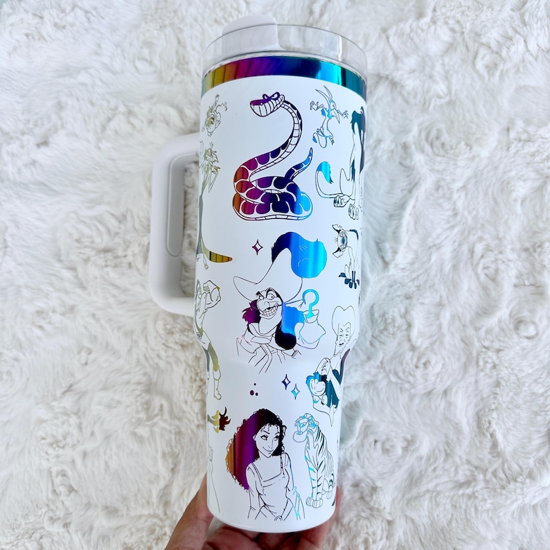 Puede incluir: Vaso blanco con asa y tapa transparente, adornado con ilustraciones de personajes de dibujos animados coloridos. El dise&ntilde;o incluye una banda iridiscente arco&iacute;ris en la parte superior. El vaso est&aacute; sostenido por una mano.