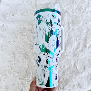 Puede incluir: Vaso blanco con asa, con un dise&ntilde;o envolvente de villanos de dibujos animados en verde, morado y azul. El vaso tiene una tapa transparente y un asa blanca. La palabra "STANLEY" es visible en el lateral.