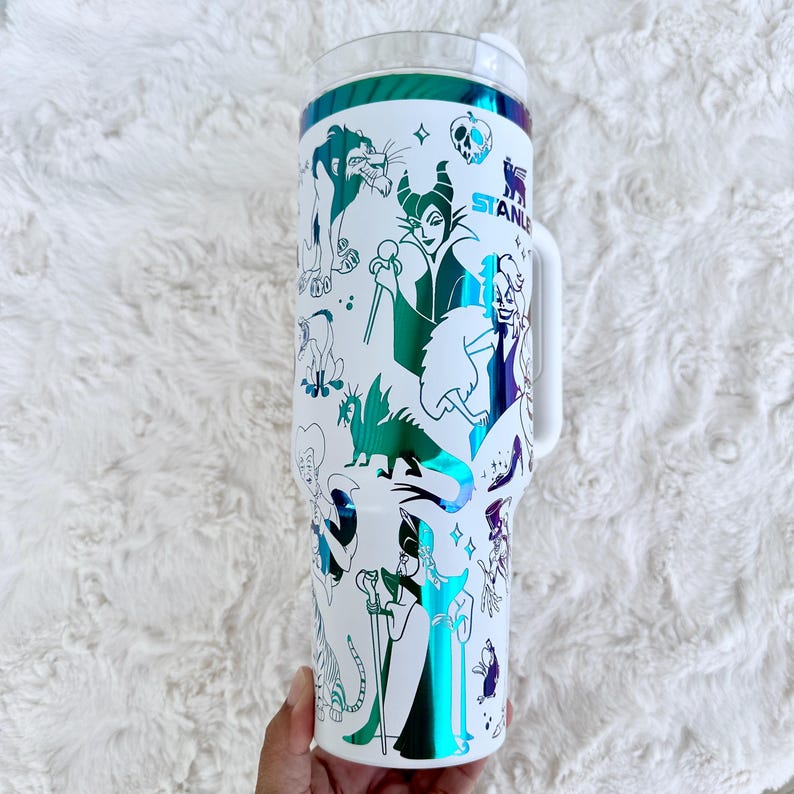 Puede incluir: Vaso blanco con asa, con un dise&ntilde;o envolvente de villanos de dibujos animados en verde, morado y azul. El vaso tiene una tapa transparente y un asa blanca. La palabra "STANLEY" es visible en el lateral.