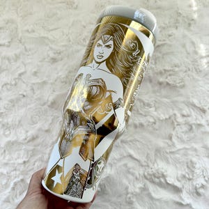 May include: White and gold tumbler featuring a Wonder Woman design. The tumbler has a gold illustration of Wonder Woman on a white background. The design includes stars and other graphic elements.