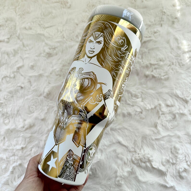 May include: White and gold tumbler featuring a Wonder Woman design. The tumbler has a gold illustration of Wonder Woman on a white background. The design includes stars and other graphic elements.