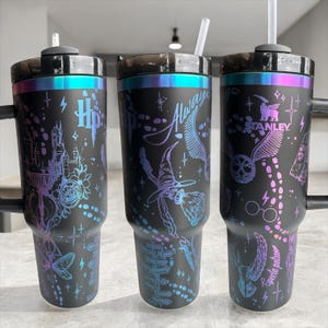 May include: Three black insulated tumblers with iridescent rims and handles. Each tumbler features a different design with Harry Potter-themed graphics, including the Hogwarts castle, a witch's hat, and the word "Always". Each tumbler has a straw.