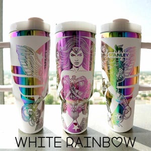 May include: Three white Stanley travel mugs with rainbow iridescent details and Wonder Woman designs. Each mug has a different design element, including the Wonder Woman logo, a portrait, and the Stanley logo. The text "WHITE RAINBOW" is at the bottom.