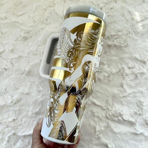May include: White travel mug with a gold and white graphic of a woman with long hair. The mug has a white handle and a grey lid. The design features gold stripes and a star.