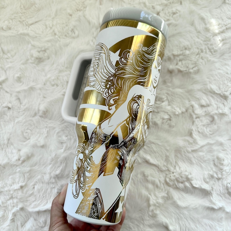 May include: White travel mug with a gold and white graphic of a woman with long hair. The mug has a white handle and a grey lid. The design features gold stripes and a star.