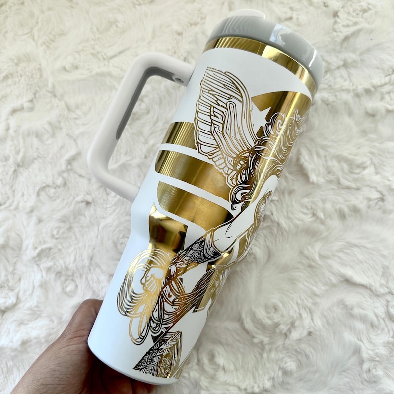 May include: White insulated tumbler with a gold and black graphic design. The tumbler features a white handle and a grey lid. The gold design includes an illustration of a winged figure. The tumbler is designed to keep beverages cold or hot.