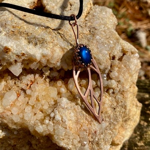 May include: A copper leaf pendant necklace with a blue iridescent gemstone. The necklace is on a black cord and is resting on a rock with white crystals.