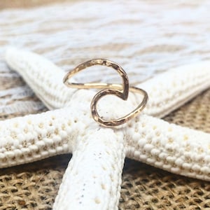 May include: A gold-colored ring with a hammered texture, shaped like a wave. The ring is displayed on a white starfish, set against a neutral background.