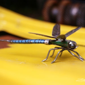May include: A metal dragonfly sculpture with blue and green accents. The dragonfly is perched on a yellow surface.