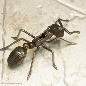 Peut inclure: Une sculpture d'une fourmi en métal réalisée à partir de matériaux recyclés, notamment une chaîne de vélo, des écrous et des boulons. La fourmi est posée sur ses pattes tendues et la tête levée.