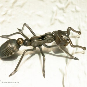 May include: A metal ant sculpture made from recycled materials, including a bike chain and a nut. The ant is silver and has a gold bead for its head.