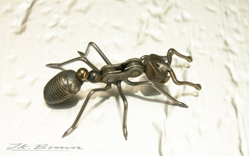 May include: A metal ant sculpture made from recycled materials, including a bike chain and a nut. The ant is silver and has a gold bead for its head.