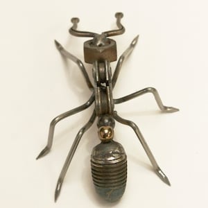 May include: A metal ant sculpture made from recycled materials. The ant is silver with a gold accent on its head. The ant is standing on a white surface.