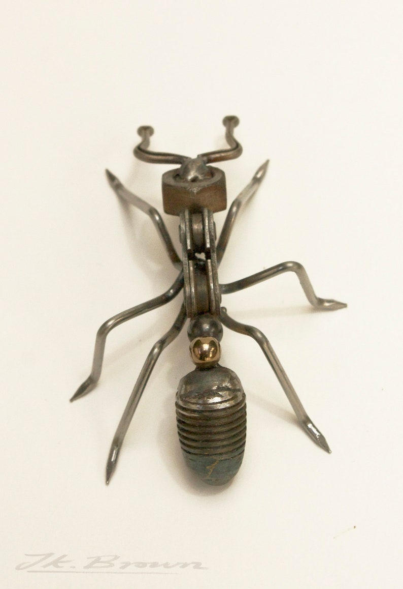 May include: A metal ant sculpture made from recycled materials. The ant is silver with a gold accent on its head. The ant is standing on a white surface.