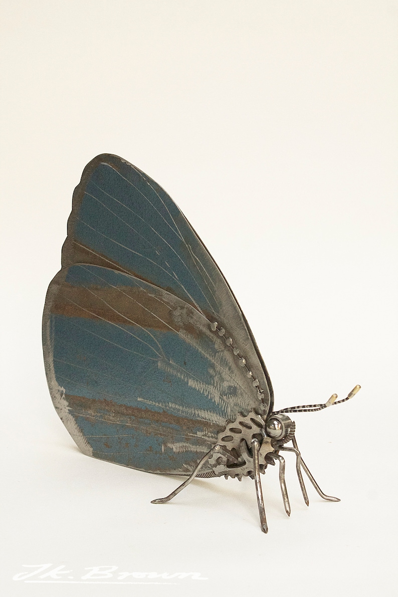 May include: A blue metal butterfly sculpture with gears and metal legs. The butterfly is made of recycled metal and has a rustic, aged look.