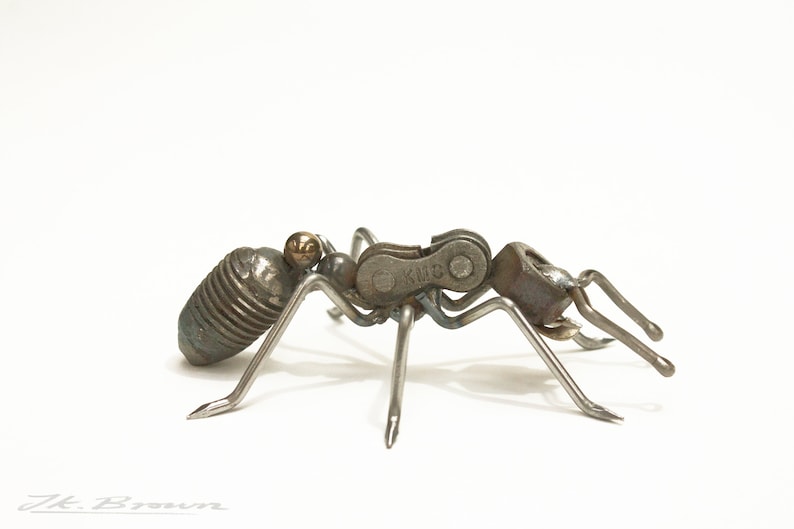 May include: A metal ant sculpture made from recycled materials, including a bicycle chain with the brand "KMC" printed on it. The ant is in a walking position with its legs extended.