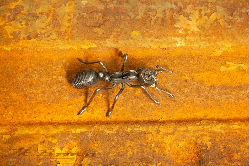 May include: A metal ant sculpture made from recycled materials, such as nuts, bolts, and chain links. The ant is posed on a rusty metal surface.