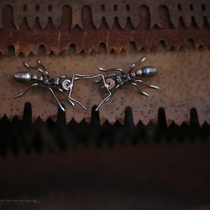 May include: Two metal ants facing each other on a rusty metal surface. The ants are made of metal and have a detailed design.