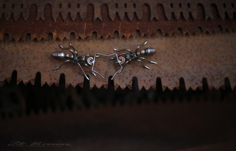 May include: Two metal ants facing each other on a rusty metal surface. The ants are made of metal and have a detailed design.
