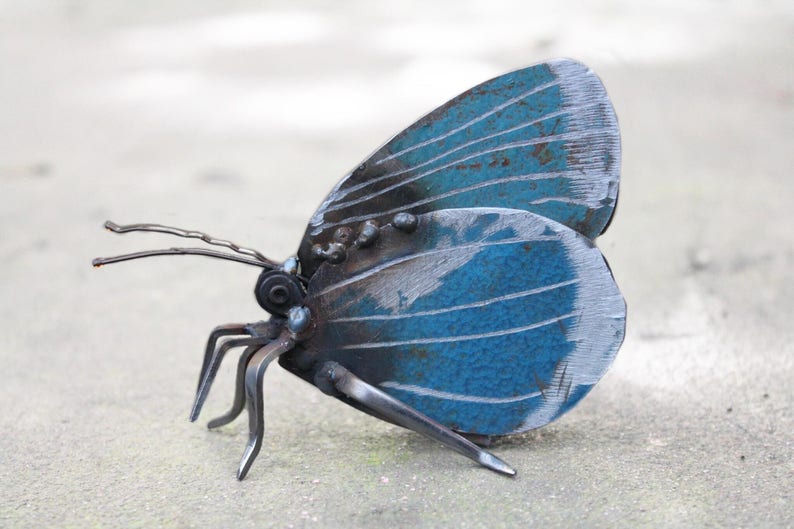 May include: A blue metal butterfly sculpture with intricate details. The wings are made of metal sheets and have a textured surface. The butterfly is perched on a gray surface.