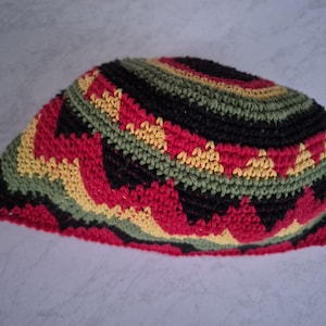 May include: A crocheted skullcap in a vibrant pattern of red, black, yellow, and green. The cap features a rounded crown and a close-knit stitch, showcasing a geometric design. The color scheme is bold and eye-catching.