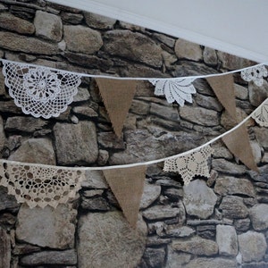 Può includere: Una ghirlanda decorativa composta da bandierine triangolari in iuta alternate a centrini in pizzo all'uncinetto bianchi e beige, infilati su un sottile nastro bianco. Gli elementi sono disposti su due file orizzontali.