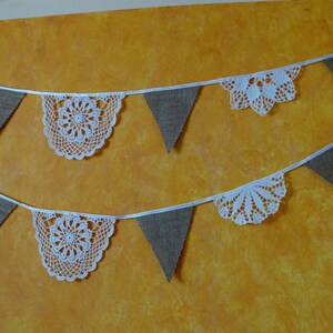 May include: A decorative bunting banner featuring alternating white crocheted lace doilies and brown triangular burlap flags suspended on a white ribbon. The garland consists of two horizontal rows of these textile elements arranged in a repeating pattern.