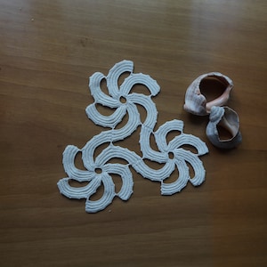 Può includere: Un centrino all'uncinetto bianco sporco fatto a mano con un motivo floreale, composto da tre motivi circolari collegati. Due conchiglie sono posizionate vicino su una superficie di legno. Il motivo e la trama intricati del centrino sono chiaramente visibili.
