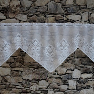 Könnte beinhalten: Weiße Spitzengardine mit Blumen- und Schleifenmuster. Die Gardine hängt vor einer Steinmauer. Die Spitze hat einen gewellten Rand und ein zartes Muster, das jedem Fenster einen Hauch von Eleganz verleiht.