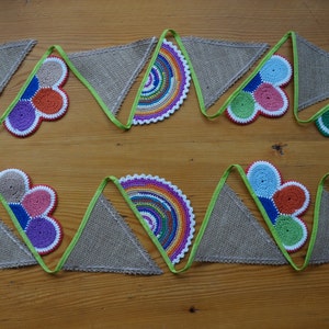 Puede incluir: Una guirnalda decorativa con banderas triangulares de arpillera que alternan con formas de ganchillo coloridas, incluyendo semicírculos arcoíris y racimos florales multicolores, unidos por un borde de cinta verde lima.