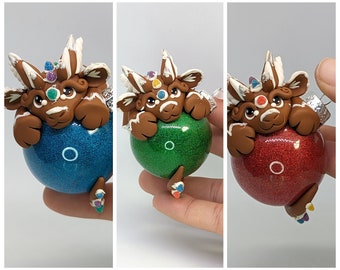 May include: Three handmade Christmas ornaments featuring a brown gingerbread dragon with white icing and colorful candy decorations. Each dragon is holding a different colored glitter ball ornament: blue, green, and red.