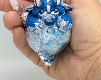 May include: Blue and white frosted dragon ornament with glitter inside. The dragon is curled up in a ball and has a white frosted finish.