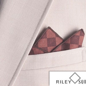 May include: A close-up of a beige suit jacket with a pocket square. The pocket square is a dark red and brown checkered pattern. The brand name "RILEY SQUARE" is visible in the bottom right corner.