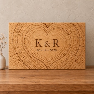 Op de afbeelding: Een rechthoekige houten plaat met een lasergegraveerde hartvorm, gevormd door natuurlijke houtnerfpatronen. In het midden staan de letters K & R boven de datum 06 • 14 • 2020 in een donker schreeflettertype op het lichte houten oppervlak.