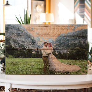 May include: A wooden wall art piece with a printed image of a couple embracing in a mountain landscape. The couple is standing in a field of green grass with a mountain range in the background. The wood grain is visible on the surface of the art piece.