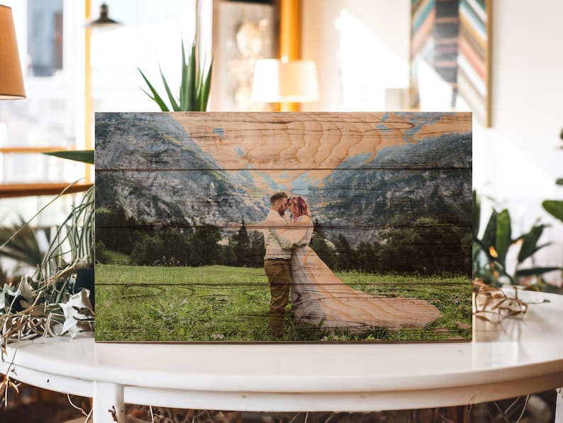 May include: A wooden wall art piece with a printed image of a couple embracing in a mountain landscape. The couple is standing in a field of green grass with a mountain range in the background. The wood grain is visible on the surface of the art piece.
