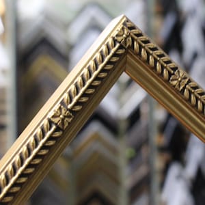 May include: Close-up of a gold picture frame corner. The frame has a decorative leaf and floral pattern along the edges. The background is blurred, showing other frames in a shop setting.