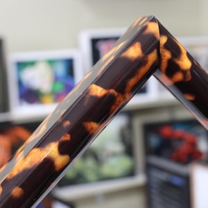 May include: Close-up of a corner of a picture frame with a tortoiseshell pattern. The frame is a dark brown color with irregular amber and yellow markings. The frame appears to be made of a glossy material.