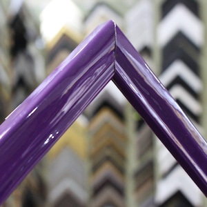 May include: Close-up of a corner of a deep purple picture frame. The frame is glossy and has a smooth finish. The frame is in focus, while the background of other frames is blurred.