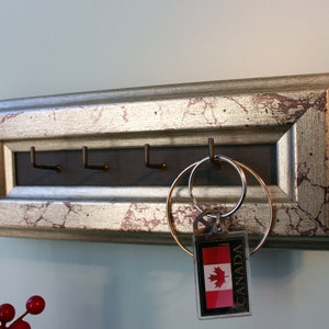 May include: A silver key holder with a distressed finish and five hooks. The key holder has a black mesh backing and a key ring with a Canadian flag keychain.